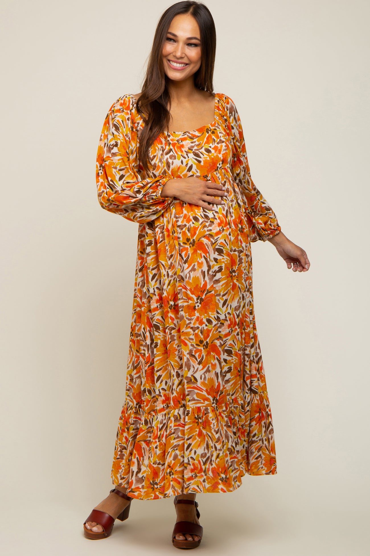 Smiling pregnant woman wearing an orange floral maternity maxi dress with long sleeves and a square neckline.