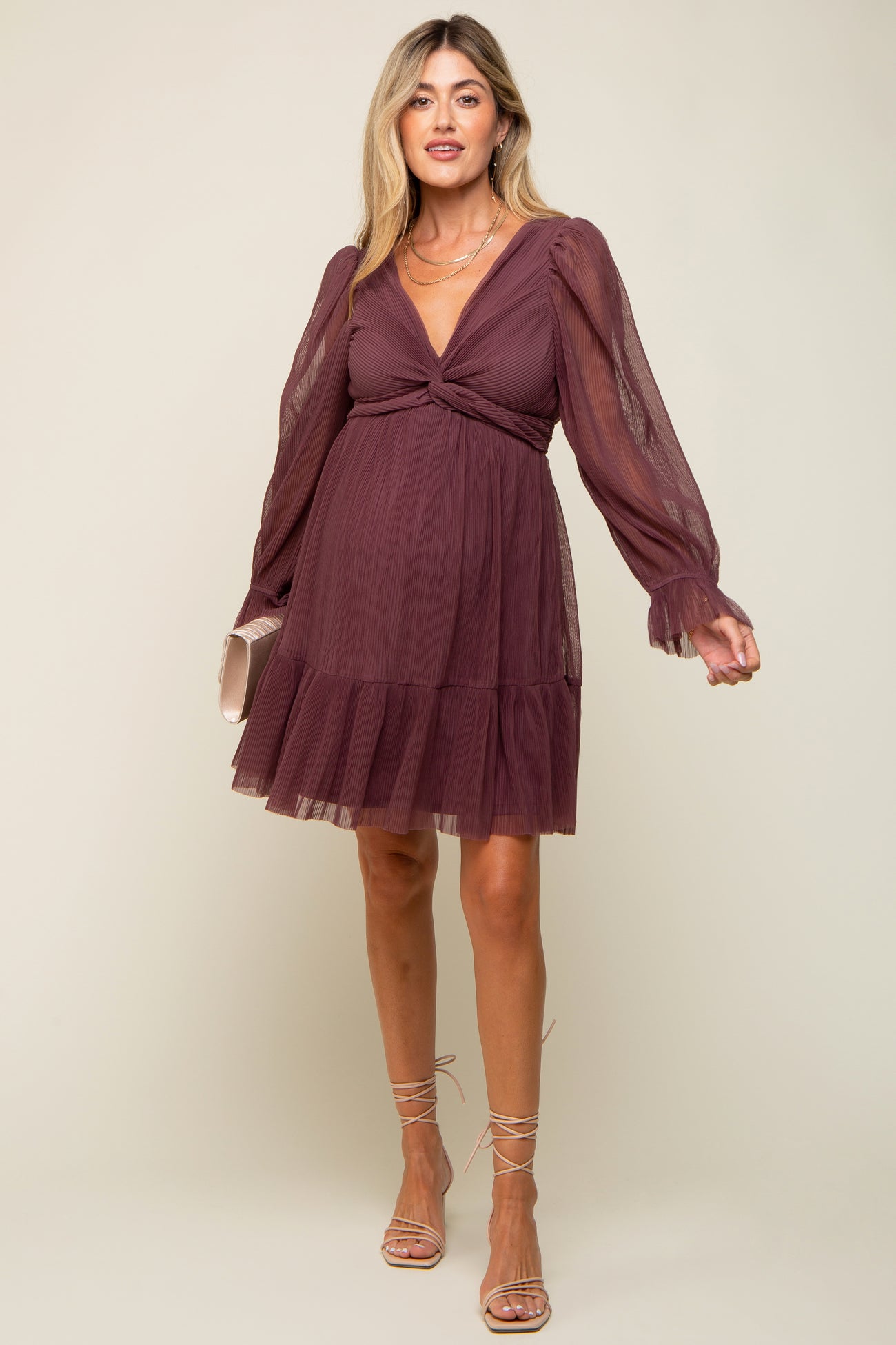 woman wearing a mauve maternity dress with long sheer sleeves and a tiered skirt, holding a small clutch