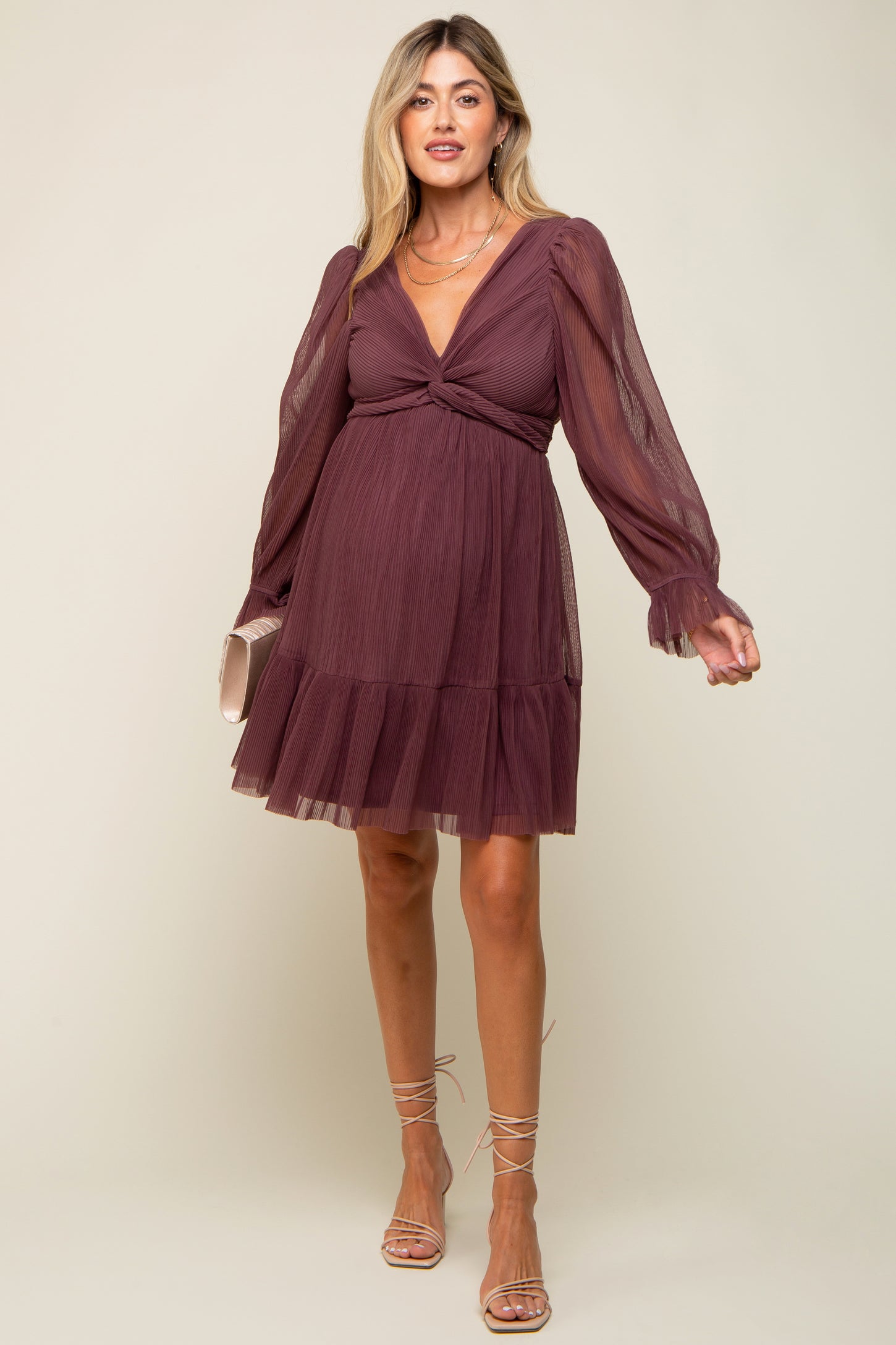 woman wearing a mauve maternity dress with long sheer sleeves and a tiered skirt, holding a small clutch