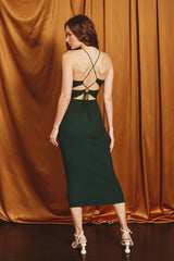 Woman in a dark green ribbed midi dress with a lace-up back and white strappy heels against gold curtains