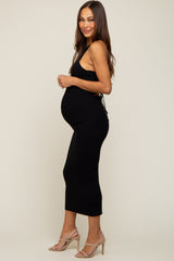 Pregnant woman wearing a black ribbed midi maternity dress and beige high heels standing against a neutral background