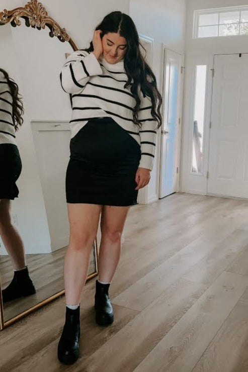 woman wearing a striped maternity sweater and a black maternity mini skirt, showing her baby bump in a mirror reflection