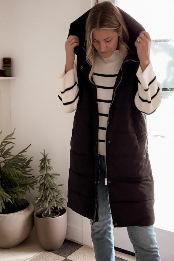 woman wearing a black puffer vest over a white and black striped sweater, standing indoors with plants in background