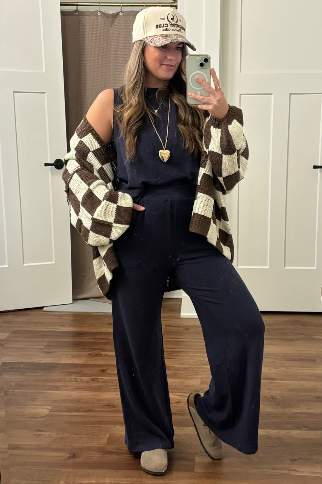 woman wearing a navy blue jumpsuit, oversized brown and cream checkered cardigan, and a beige baseball cap, posing in front of a mirror