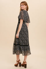 woman wearing a black floral midi dress with short sleeves and a tiered hem, standing with her back to the camera