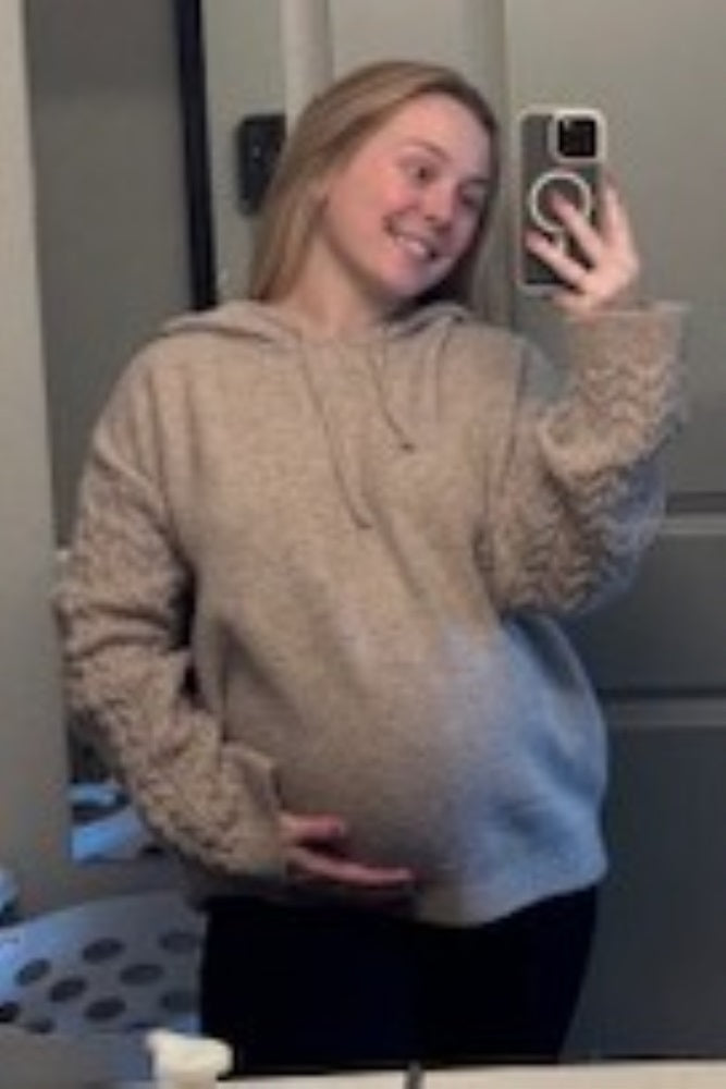 woman wearing a beige maternity hoodie with textured sleeves and holding her baby bump in a bathroom mirror