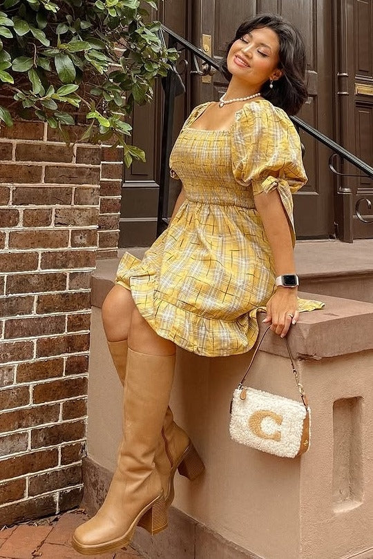 woman in yellow plaid puff sleeve dress and tan knee high boots sitting on a stone ledge