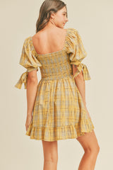 woman wearing a yellow plaid short dress with puff sleeves and a fitted bodice, showing a ruffled hem and tie details