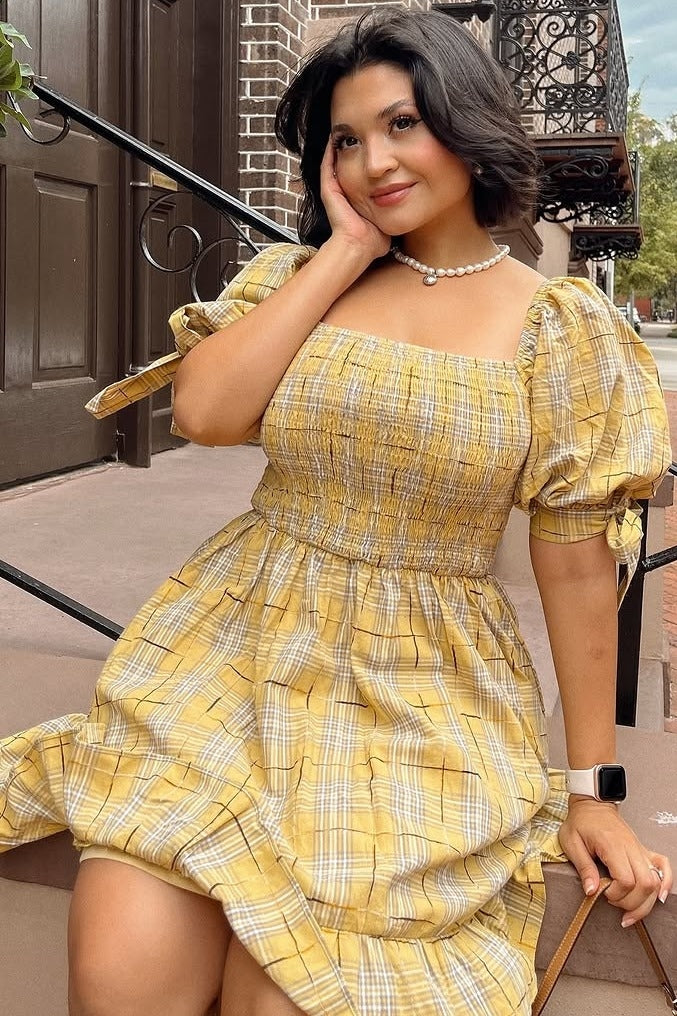 woman wearing a yellow plaid puff sleeve dress with smocked bodice sitting on steps and resting her hand on her face