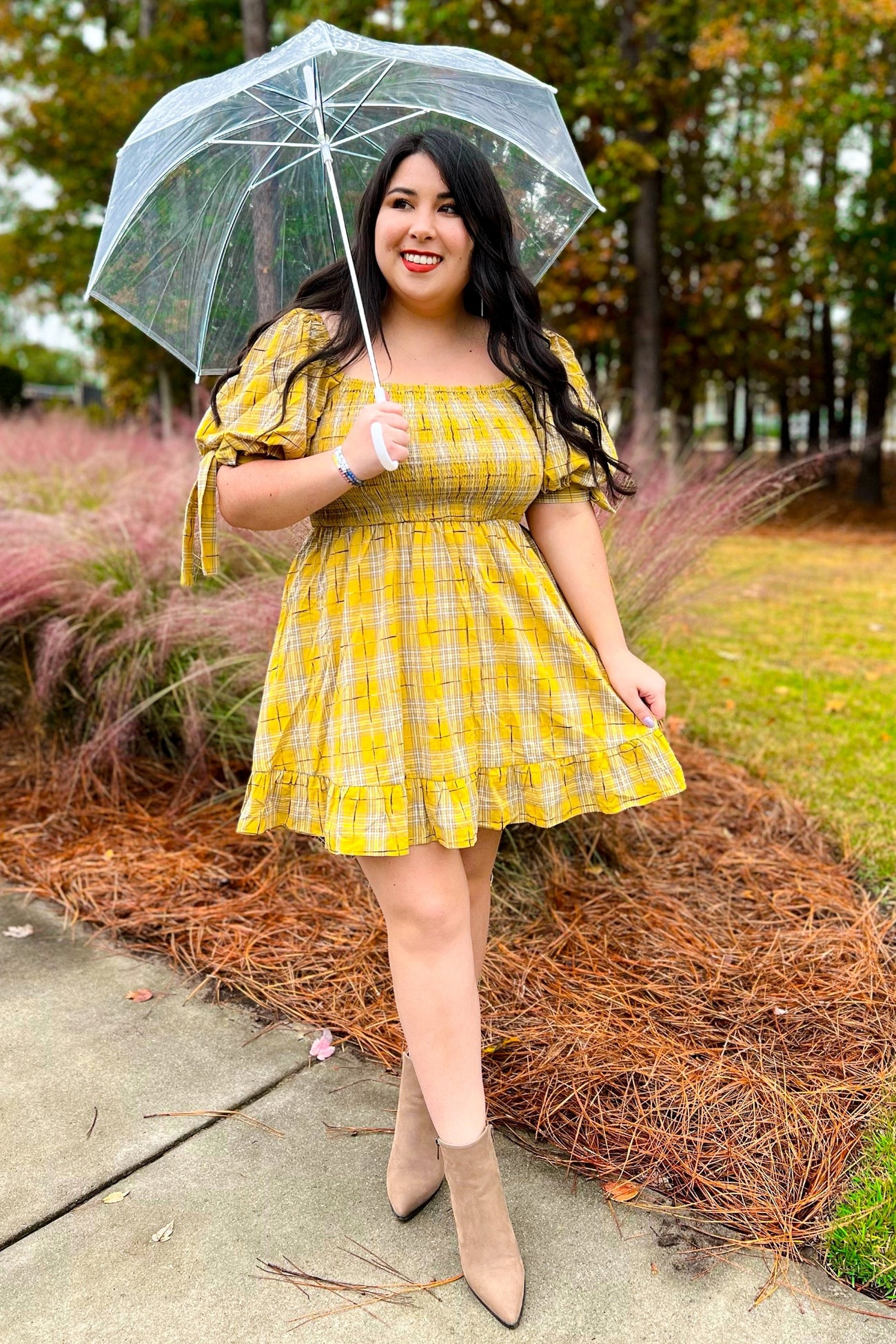 woman wearing a yellow plaid off-shoulder dress and holding a clear umbrella in an outdoor setting