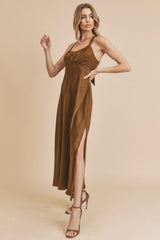 woman wearing a brown sleeveless maxi dress with a side slit and thin straps, posing with one hand on her hip