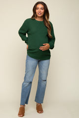 woman wearing a green knit maternity sweater and distressed blue jeans, holding her baby bump