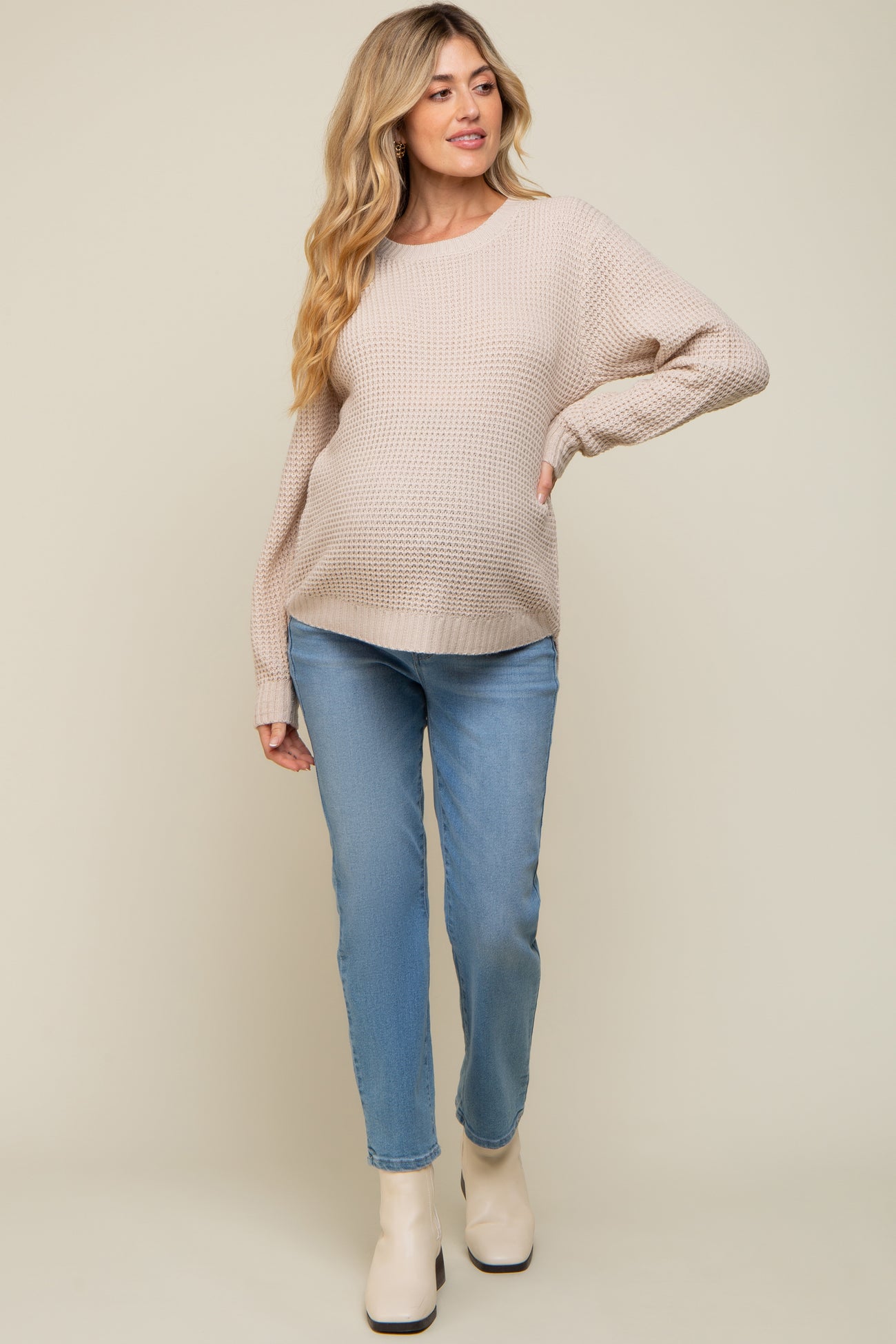 woman wearing a beige knitted maternity sweater and light blue jeans, showing off her baby bump