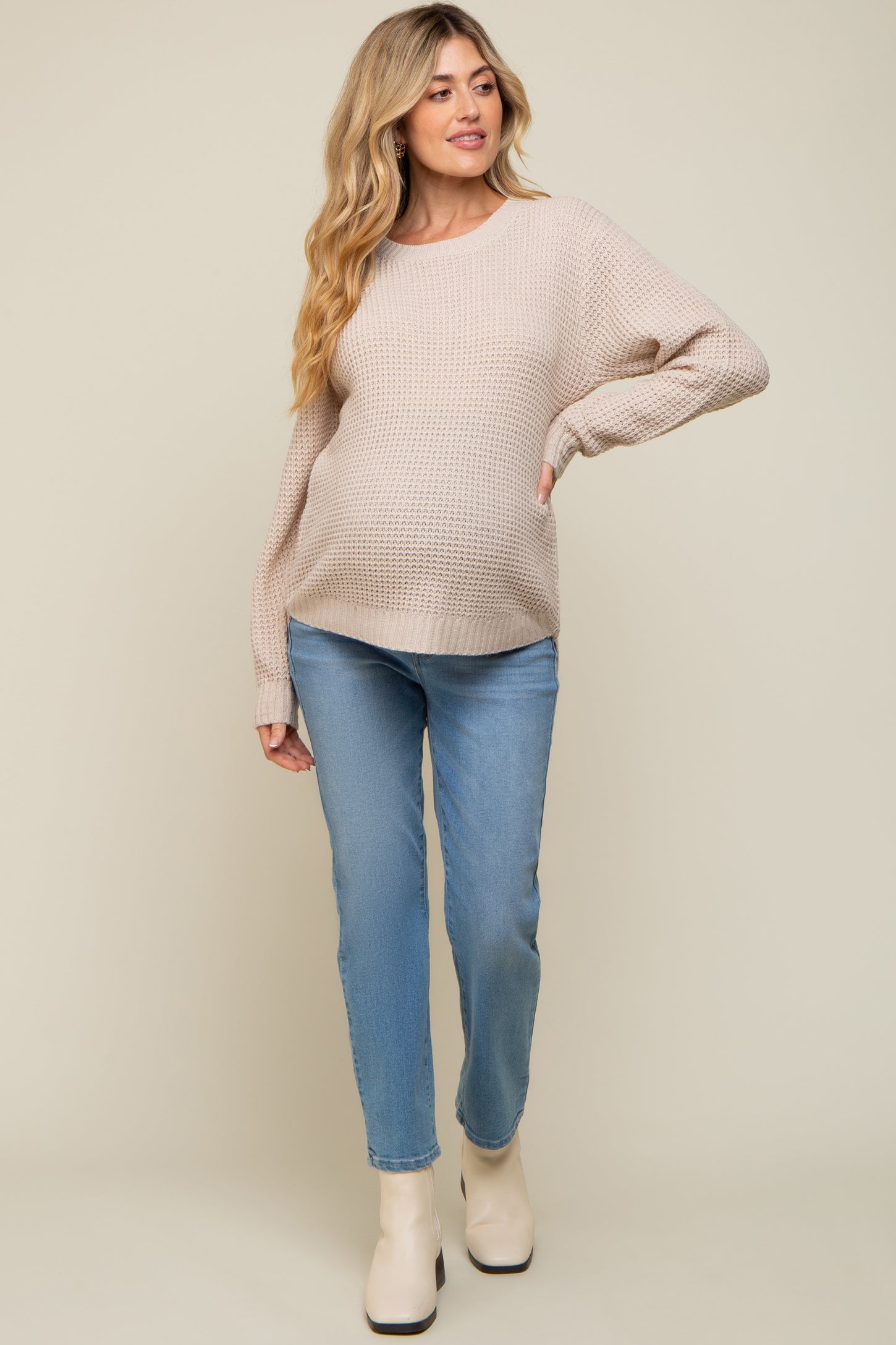 woman wearing a beige knitted maternity sweater and light blue jeans, showing off her baby bump
