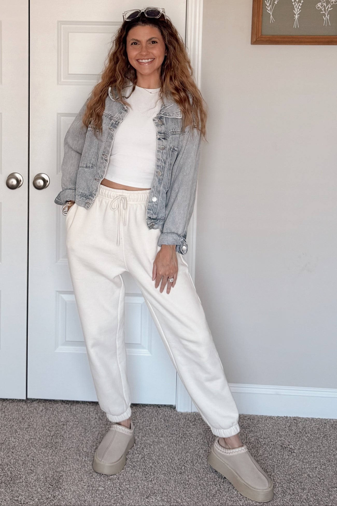 woman wearing a white cropped top, gray denim jacket, cream joggers, and beige slip-on shoes with curly hair styled down
