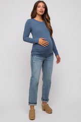 woman wearing a blue long sleeve maternity top and holding her baby bump, paired with distressed jeans and clogs