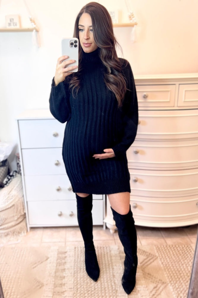 woman wearing a black ribbed maternity sweater dress and holding her baby bump while posing in front of a mirror