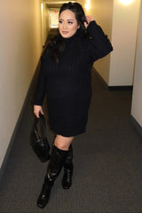 woman wearing a black ribbed maternity sweater dress and knee-high black boots, posing in a hallway