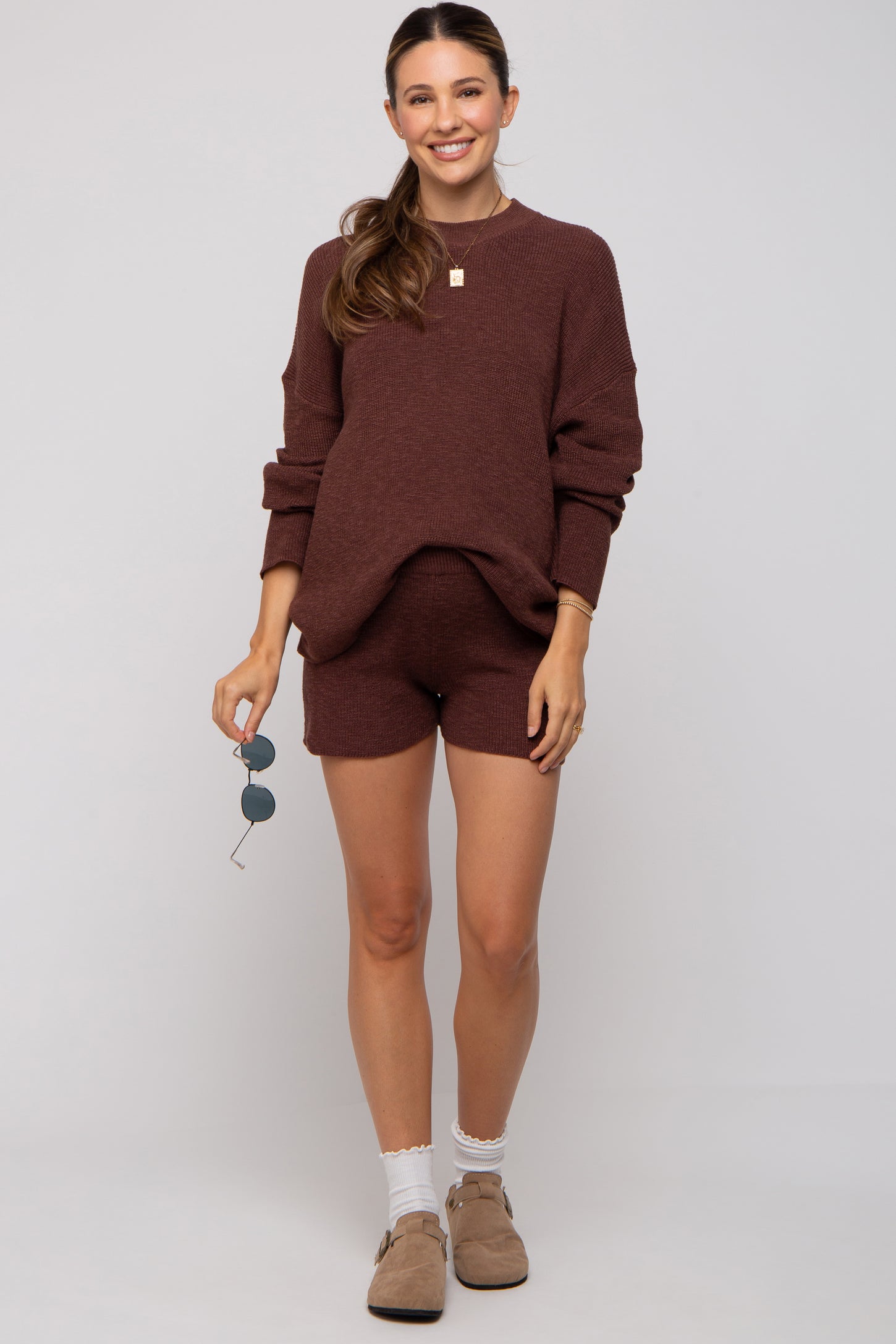 woman wearing a maroon maternity sweater and matching shorts, showcasing her baby bump, styled with sunglasses and light shoes