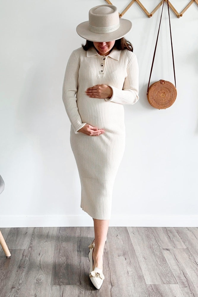 woman wearing a cream ribbed midi maternity dress with buttons, holding her baby bump, and wearing a wide-brimmed hat