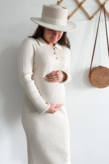 woman wearing a cream ribbed maternity dress with button details and holding her baby bump, accessorized with a wide-brim hat