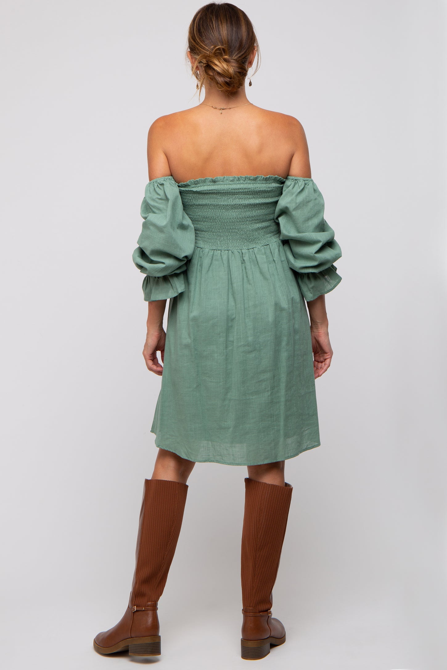 woman wearing a green off-the-shoulder dress with puff sleeves and knee-high brown boots, viewed from the back