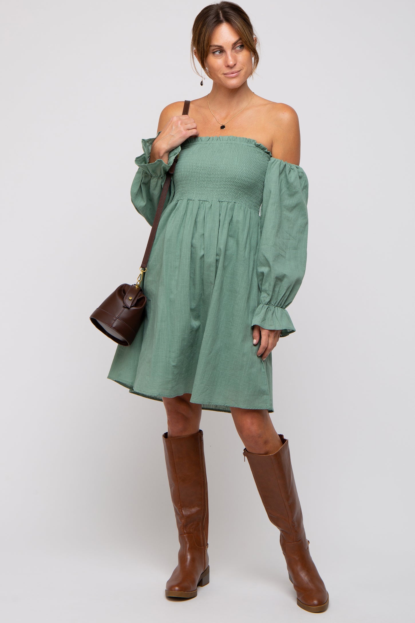 woman wearing a green off-shoulder smocked mini dress with long puff sleeves and brown knee-high boots, holding a brown handbag