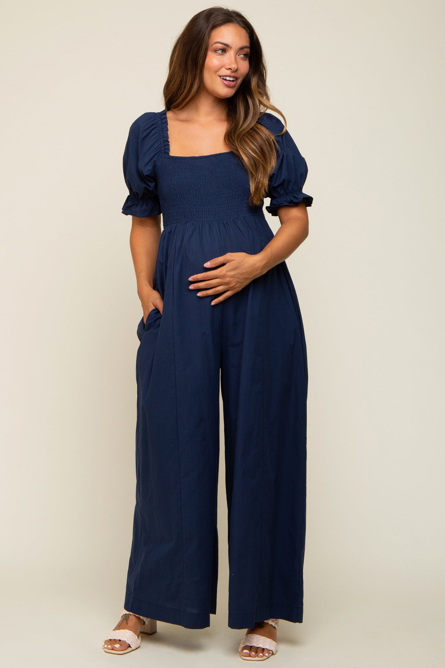 woman wearing a navy blue smocked maternity jumpsuit with puff sleeves, holding her baby bump