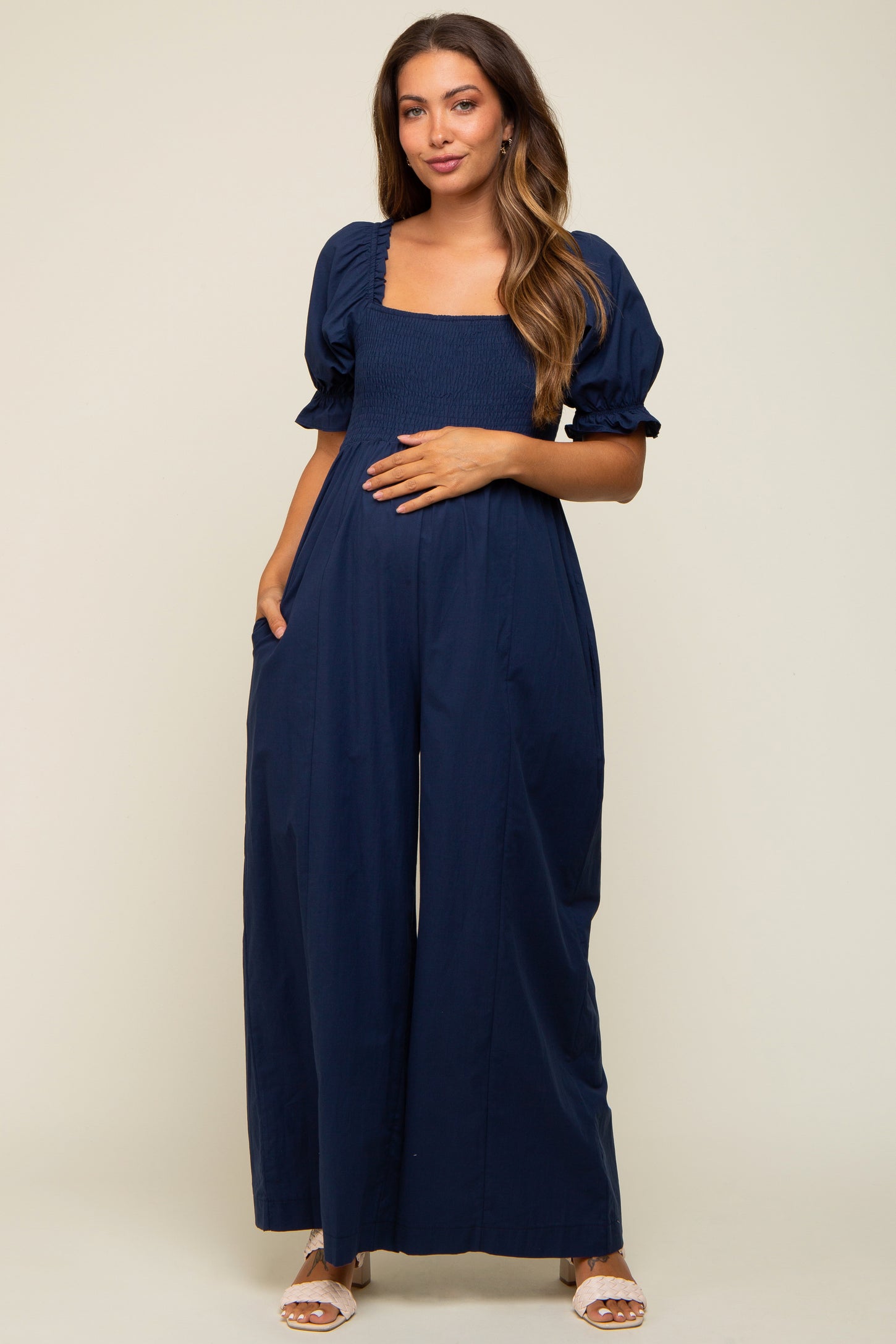 woman wearing a navy blue maternity jumpsuit with puff sleeves, elastic smocked bodice, and side pockets, showing her baby bump