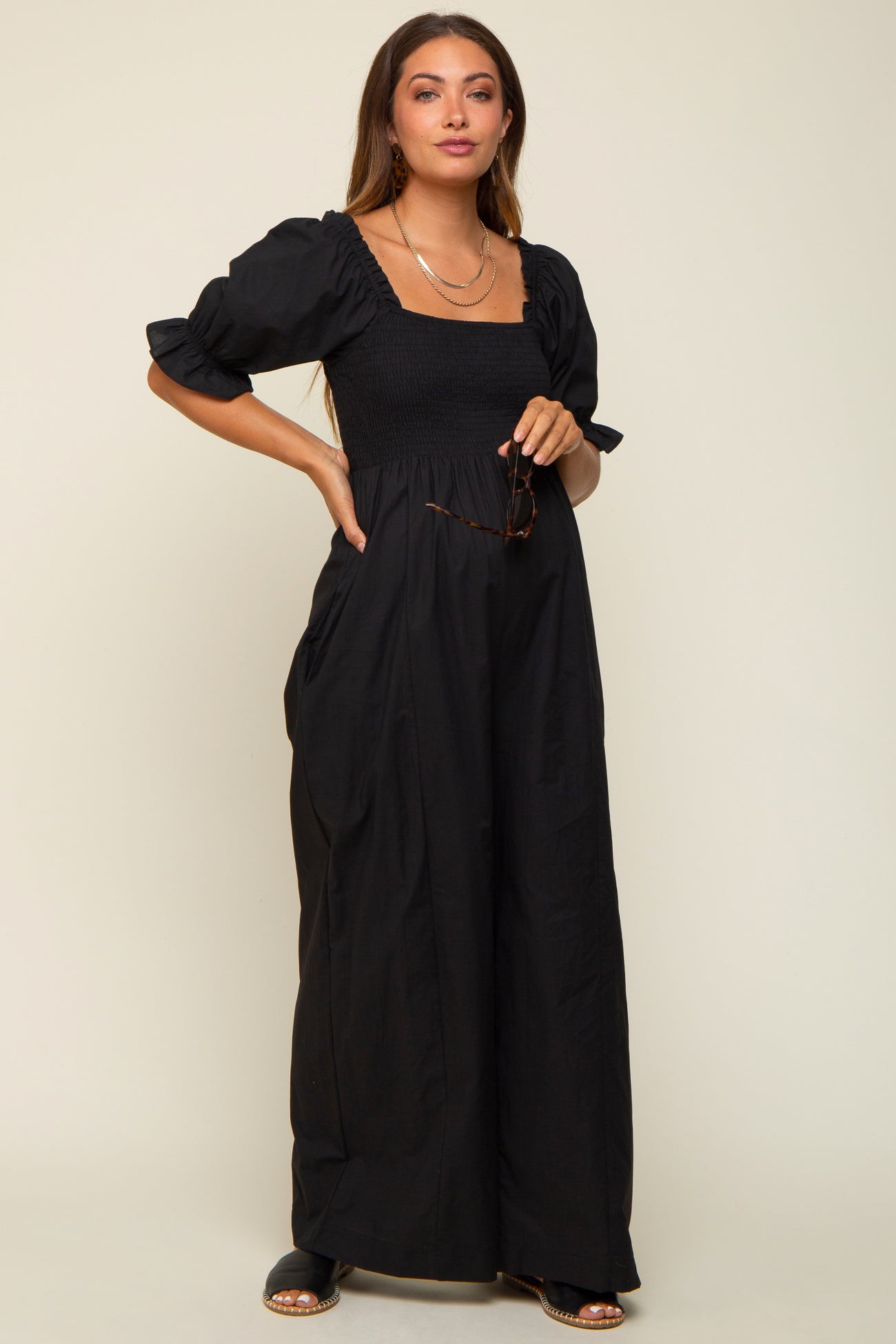 woman wearing a black maternity maxi dress with puff sleeves and a smocked bodice, holding sunglasses and showcasing her baby bump