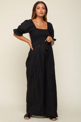 woman wearing a black maternity maxi dress with puff sleeves and a smocked bodice, holding sunglasses and showcasing her baby bump
