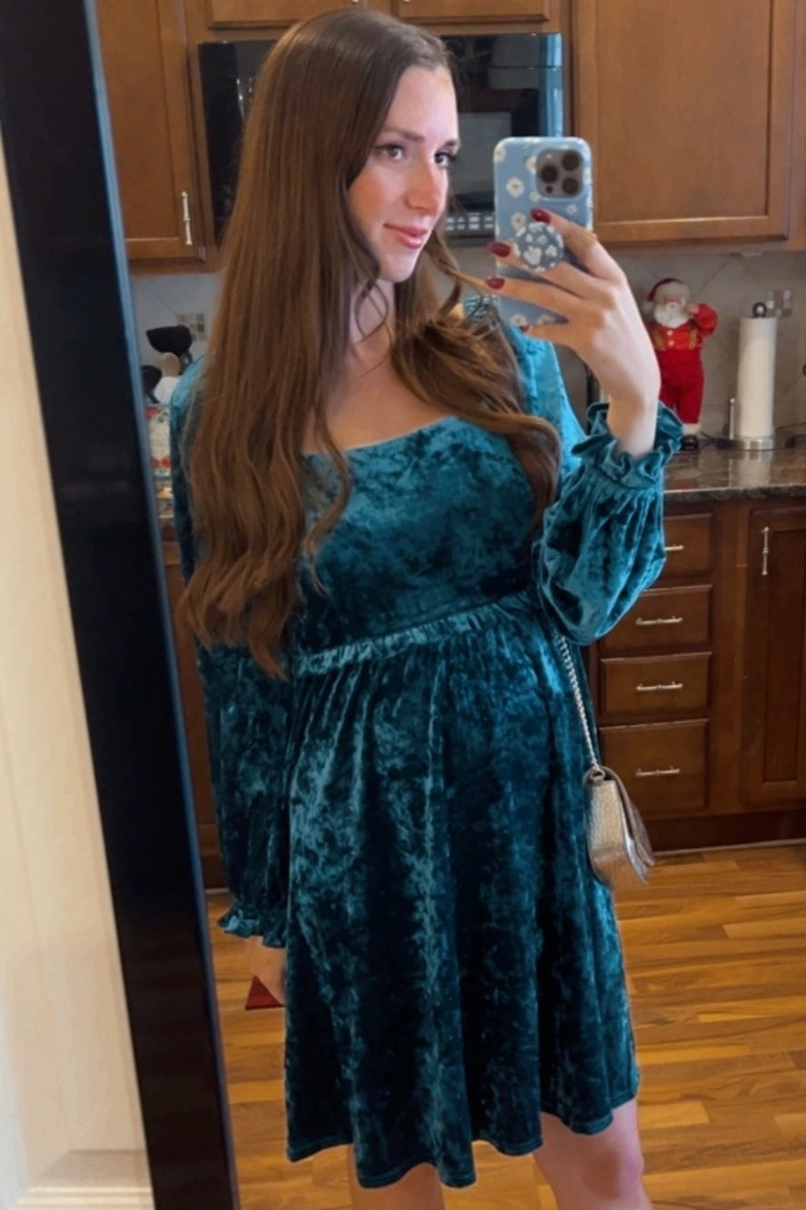 woman wearing a teal velvet long sleeve dress with a fitted bodice and flared skirt, posing in front of a mirror