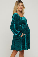 woman wearing a teal velvet long sleeve maternity dress with a fitted waist and pockets, showcasing her baby bump