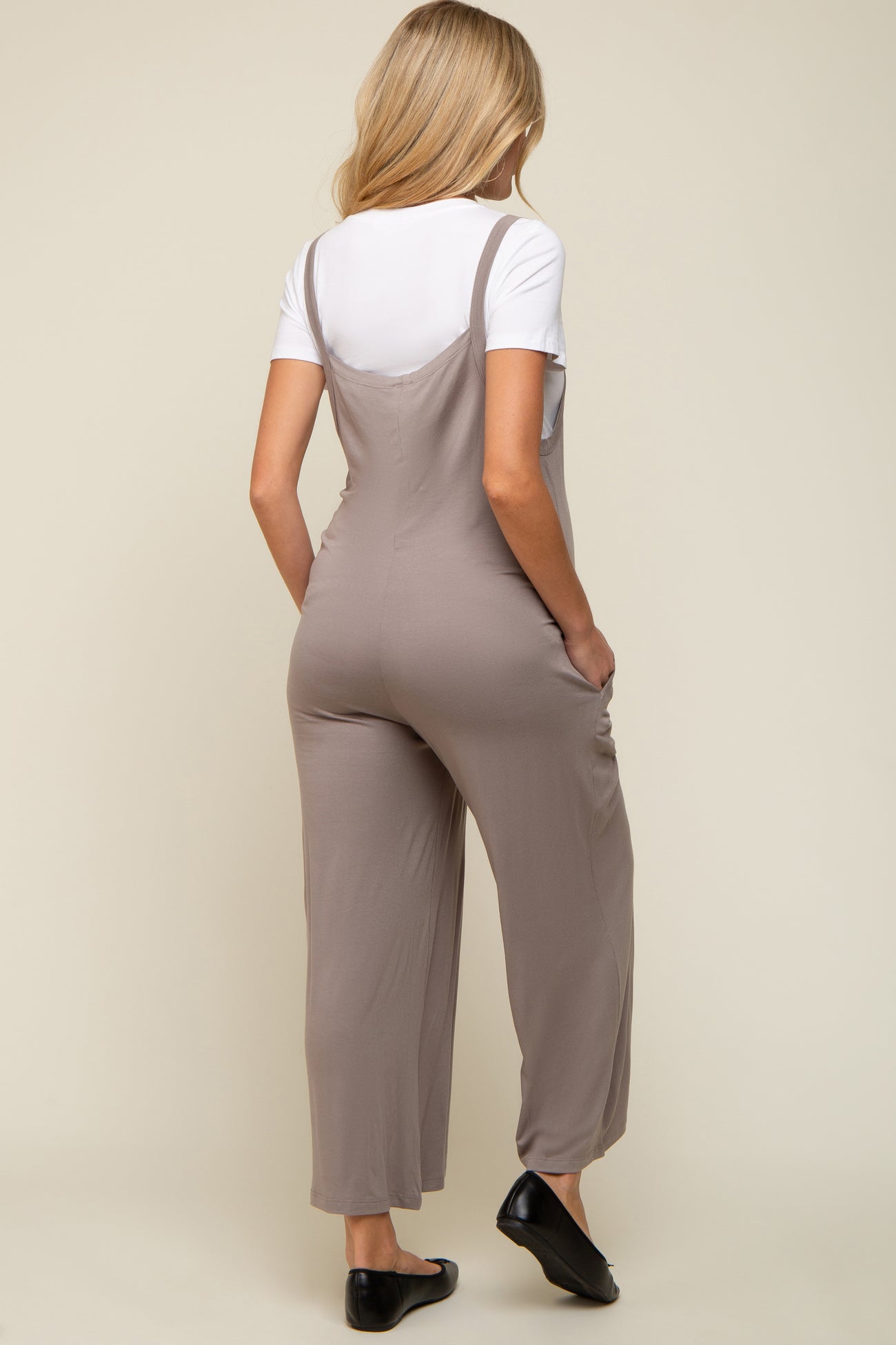 woman wearing a light gray maternity jumpsuit with adjustable straps and side pockets, viewed from the back