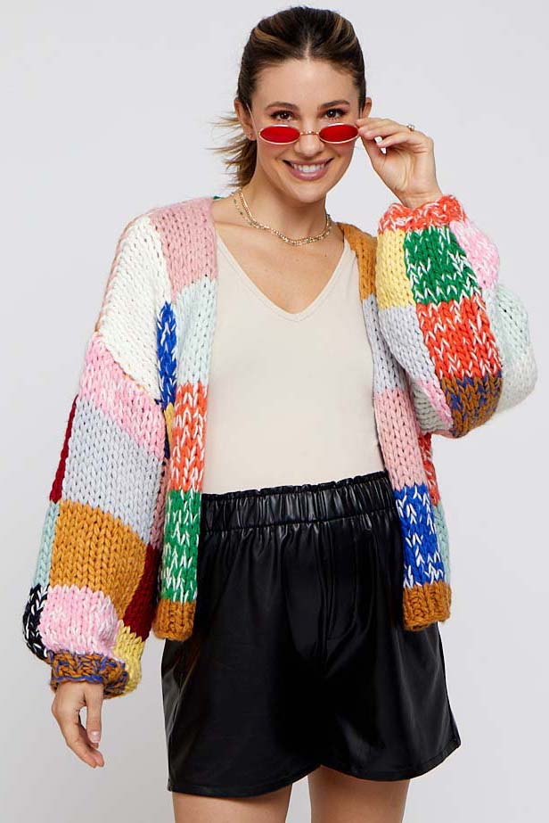 woman wearing a colorful knitted cardigan over a beige v-neck top and black faux leather shorts, smiling and holding red sunglasses