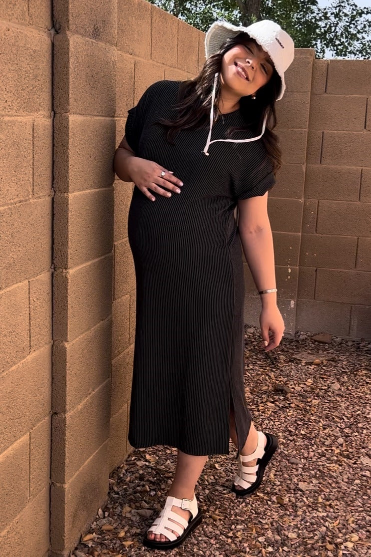 woman wearing a black striped maternity dress with a side slit, holding her baby bump and wearing a white bucket hat