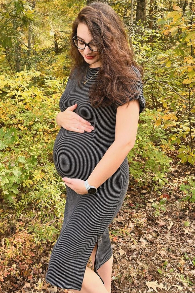 woman wearing a gray ribbed maternity dress, holding her baby bump in an outdoor setting with autumn foliage