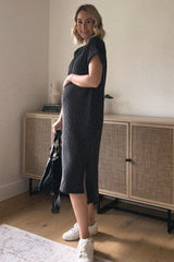 woman wearing a dark gray ribbed maternity dress with side slits and holding her baby bump, styled with a black bag and sneakers
