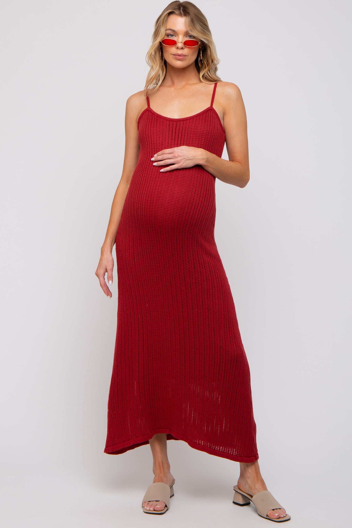 woman wearing a red ribbed maternity maxi dress and holding her baby bump, paired with red sunglasses and tan sandals
