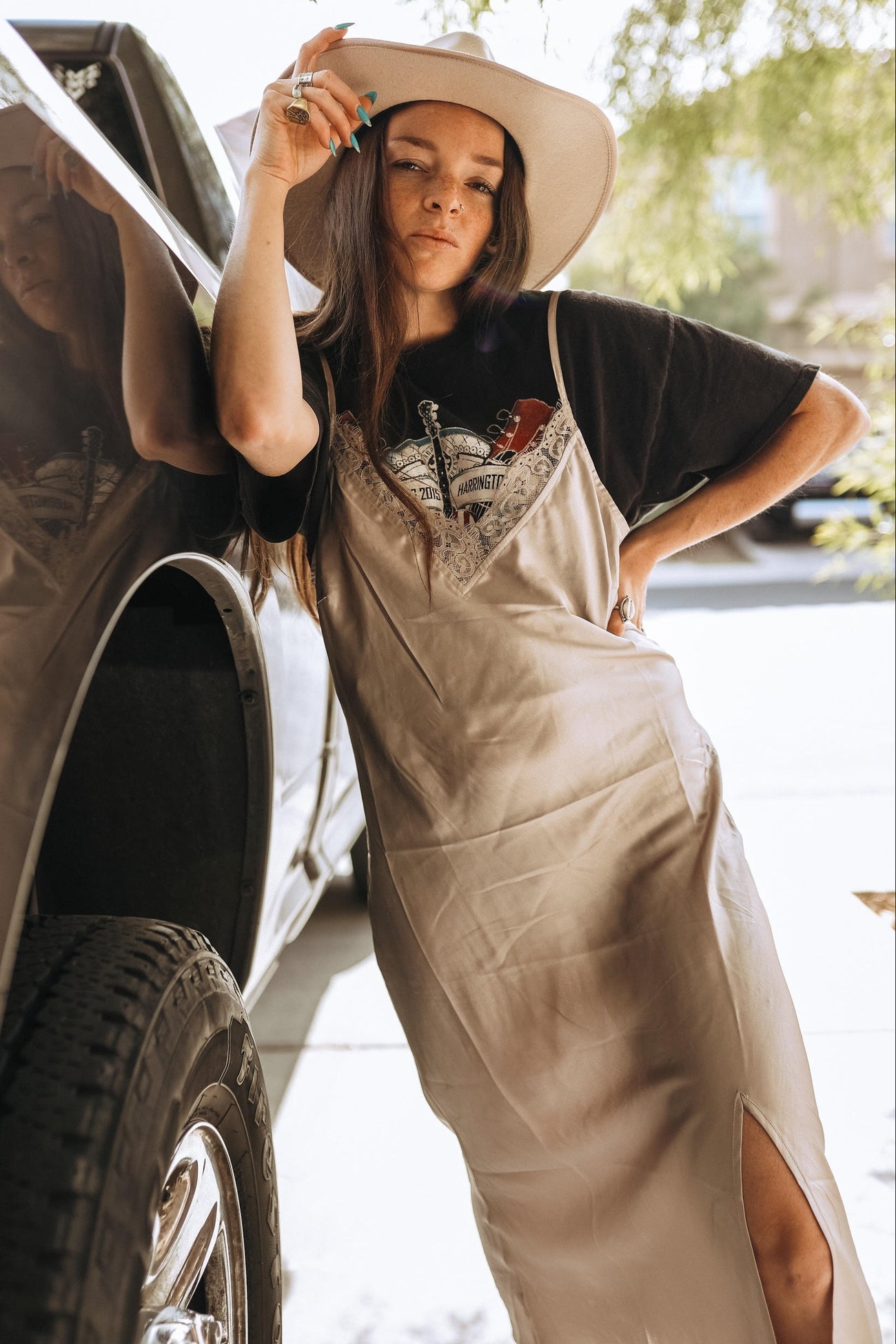 woman wearing a beige slip dress over a black t-shirt, posing next to a truck with a wide-brimmed hat