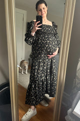 woman wearing a black floral maternity maxi dress and holding her baby bump with a dog in the background