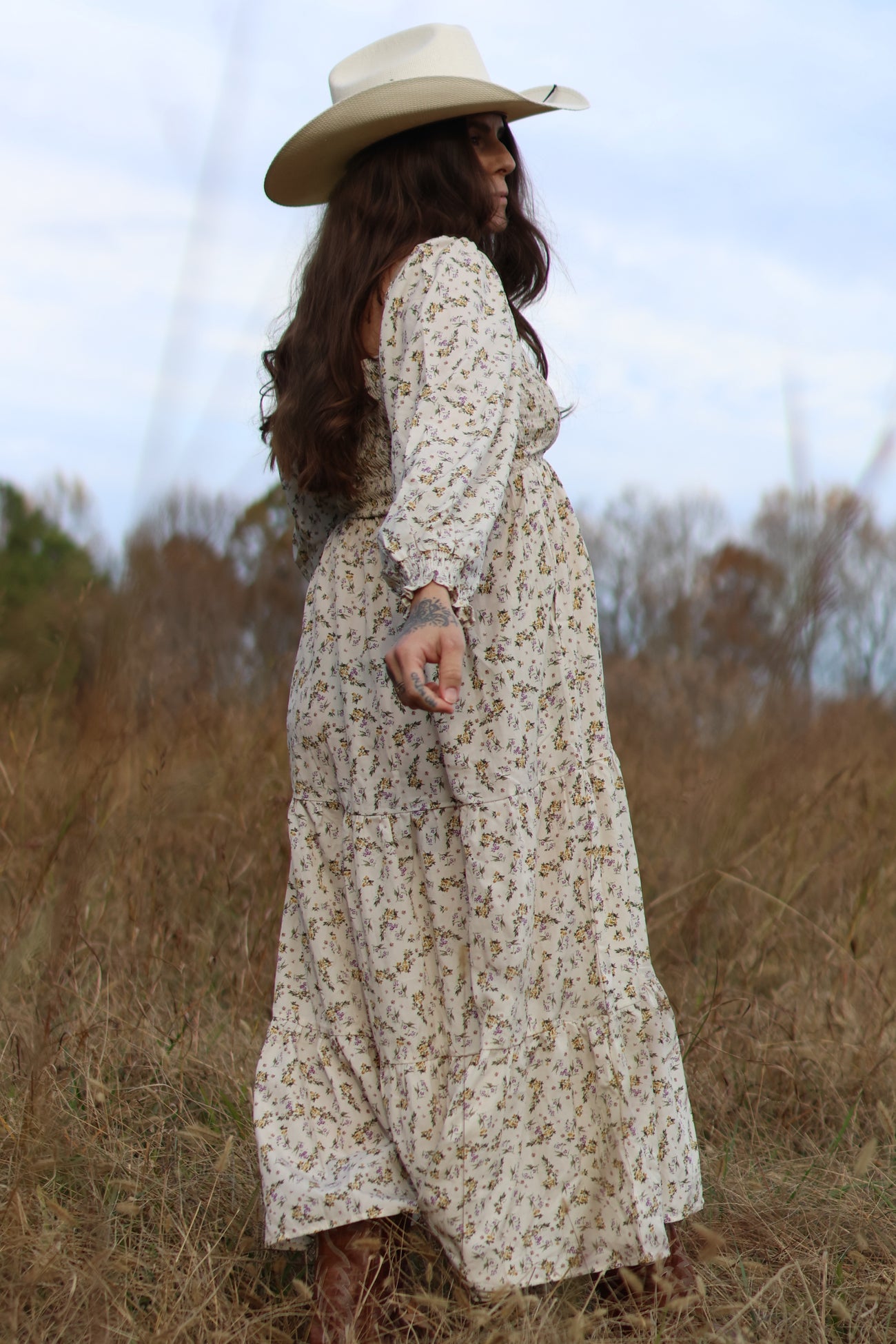 woman wearing a floral print maternity maxi dress and a wide-brimmed hat, standing in a field with a visible baby bump