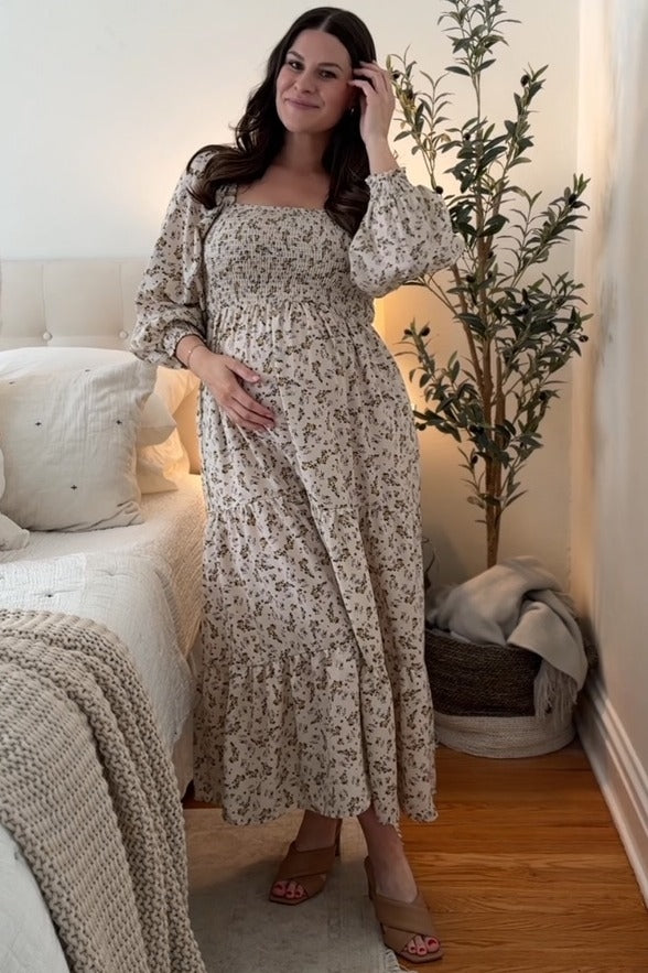 woman wearing a beige floral maternity maxi dress, holding her baby bump while posing in a cozy bedroom setting
