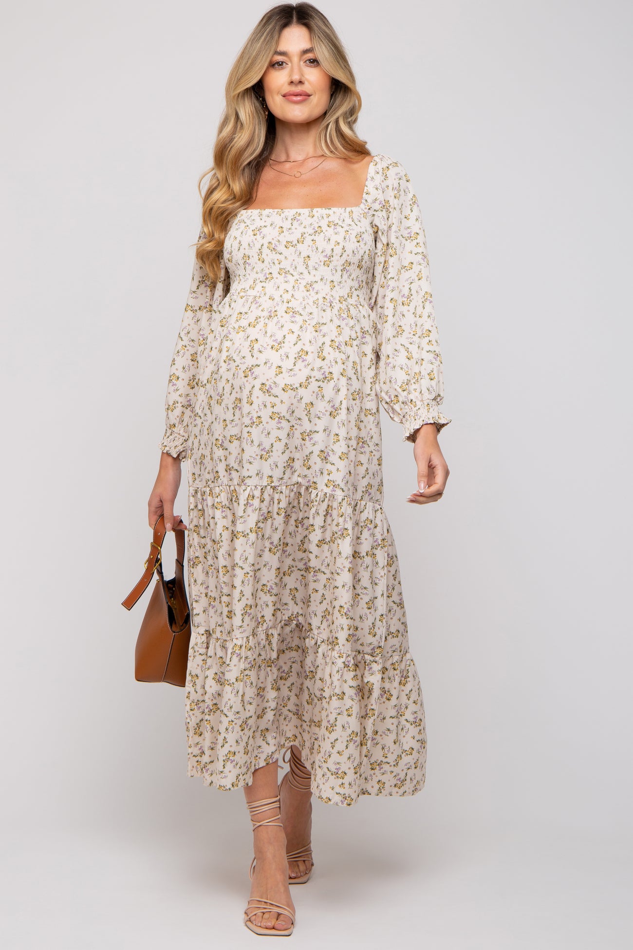 woman wearing a light floral maternity dress with a square neckline and tiered skirt, holding a brown handbag and showing baby bump