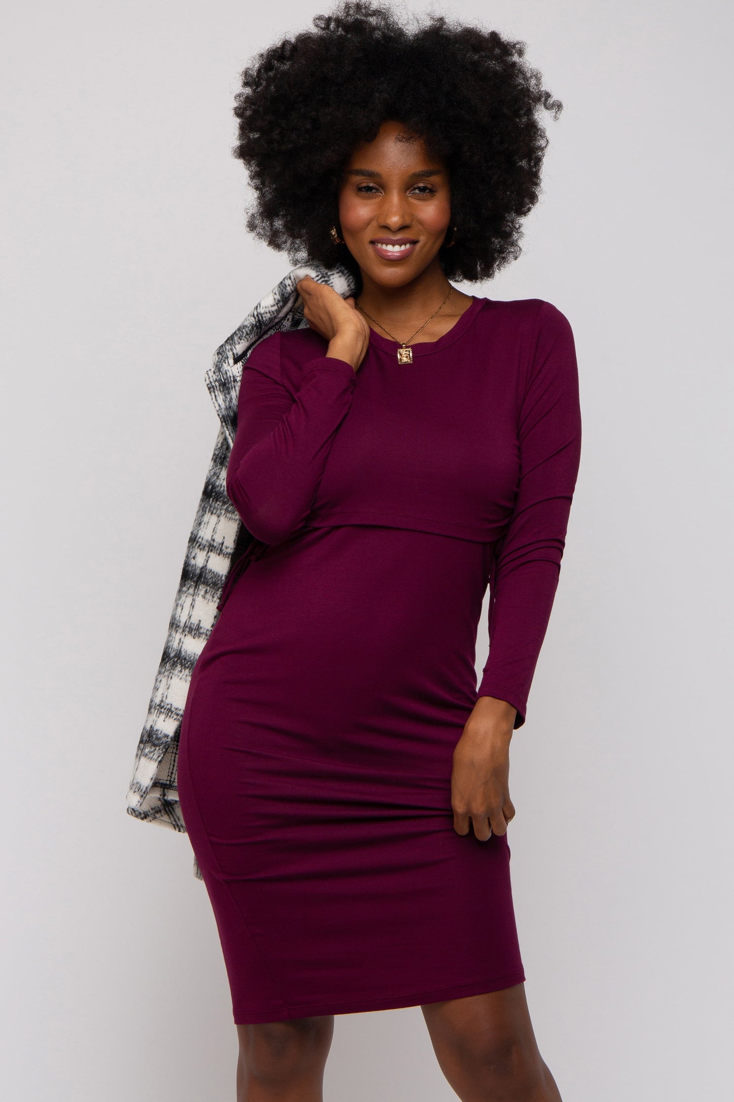 woman wearing a fitted burgundy long sleeve dress with a round neckline and posing with a black and white blazer over her shoulder