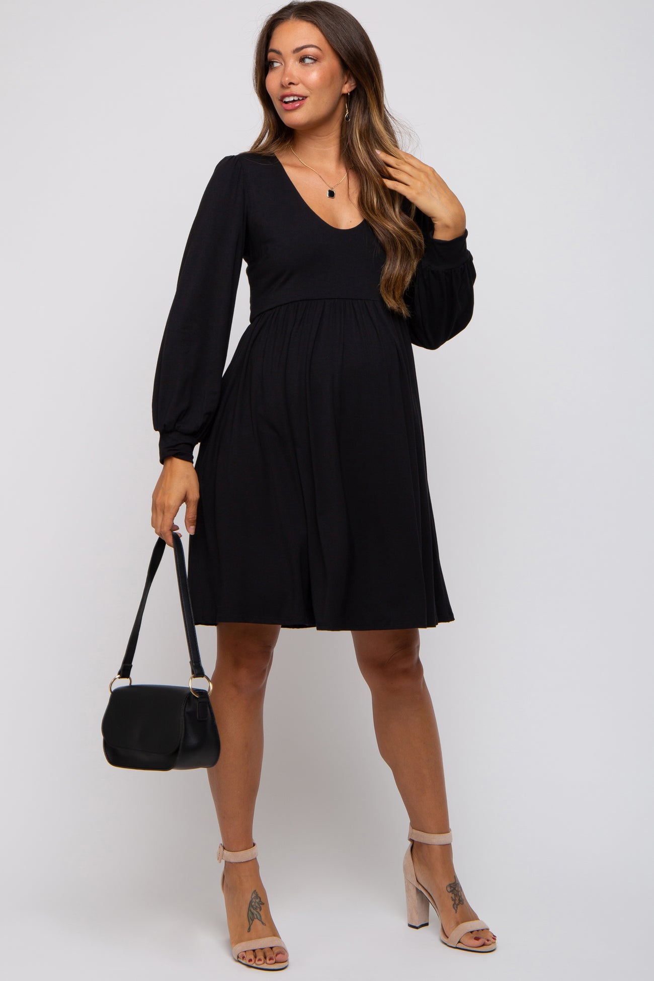 woman wearing a black maternity dress with a v-neck and long sleeves, holding a black handbag and smiling