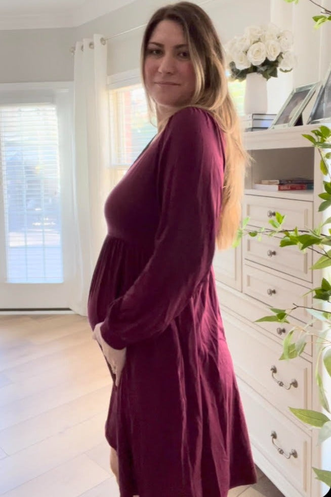 woman wearing a burgundy long sleeve maternity dress, gently holding her baby bump while standing indoors