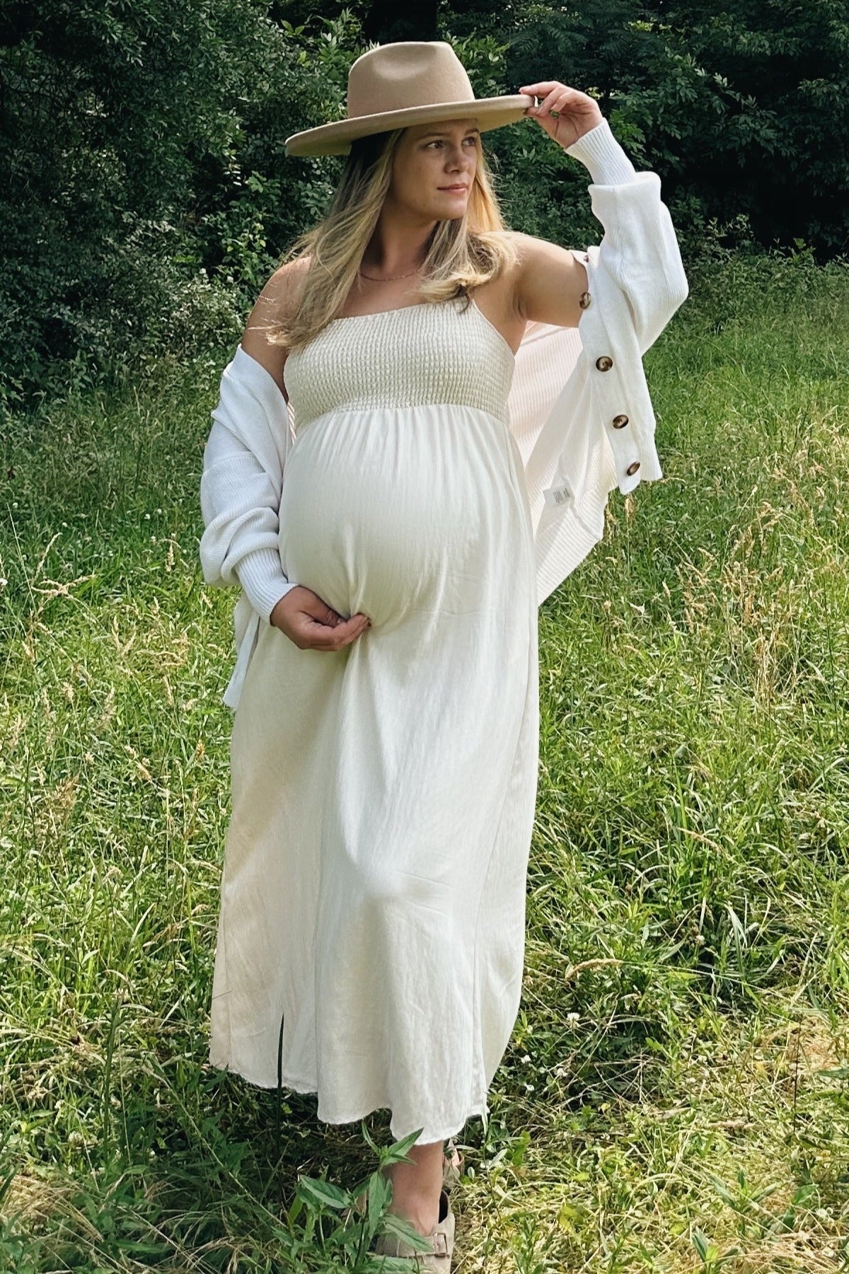 woman wearing a white maternity maxi dress with a knitted bodice, holding her baby bump and wearing a tan hat