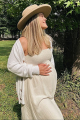 woman wearing a white strapless maternity dress and a cream cardigan, holding her baby bump outdoors
