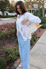 woman wearing a white cardigan and light blue wide-leg jeans, holding a cream handbag, smiling outdoors near flowers